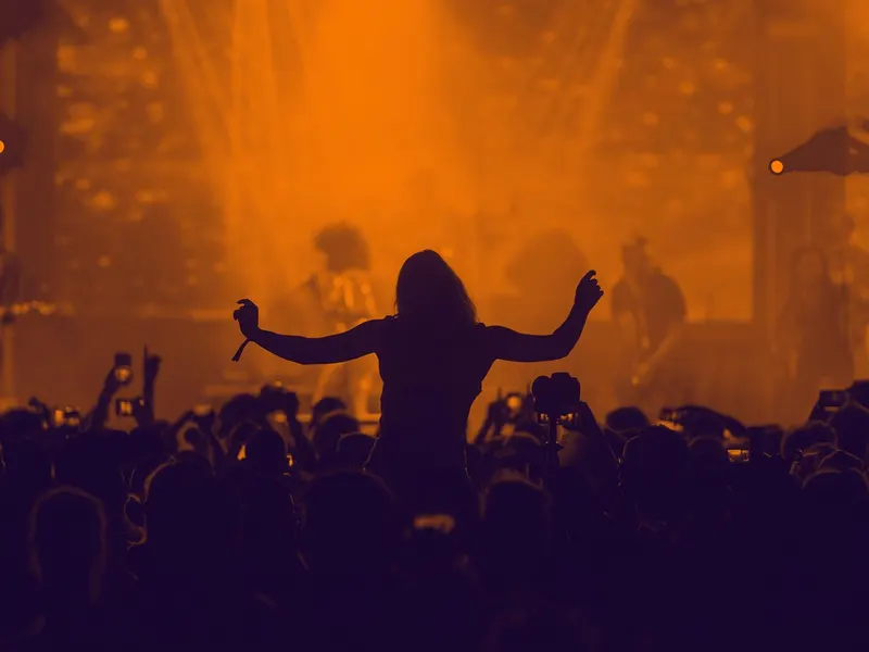 Silhouetted fan with arms raised at an orange-lit concert crowd filming on phones
