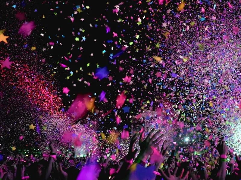 Crowd with raised hands under falling pink and purple confetti at a concert