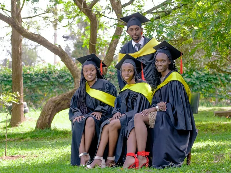 Four graduates in black gowns and mortarboards with yellow sashes sit on grass outdoors